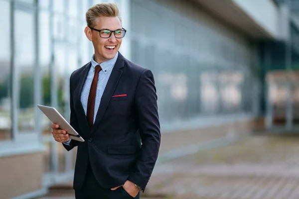 그의 PC 태블릿을 사용하여 우아한 미소 젊은 사업가. 사무실 건물 앞에서 태블릿 PC가있는 트렌디 한 옷을 입은 남성 임원의 초상화. 현대 기술 개념 스톡 사진