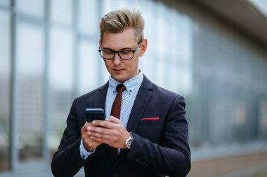 Mutlu işadamı, ofisin önünde duran bir mesaj okurken gülümsüyor. Açık havada akıllı telefon kullanarak trendy giysiler içinde Erkek icra yöneticisi. Modern teknoloji kavramı
