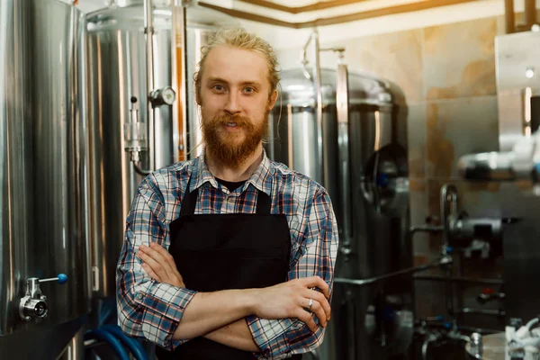 Un ritratto di un bel birraio con i dreadlocks in uniforme alla fabbricazione della birra con contenitori di metallo sullo sfondo, che sta facendo la birra sul suo posto di lavoro nel birrificio . — Foto Stock