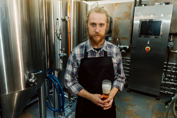 Un ritratto di un bel birraio con i dreadlocks in uniforme alla fabbricazione della birra con contenitori di metallo sullo sfondo, che sta facendo la birra sul suo posto di lavoro nel birrificio . — Foto Stock