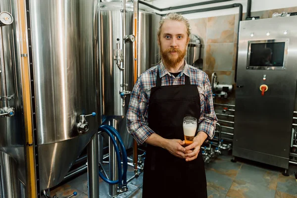 Portrét hezkého pivovaru s příšerami v uniformě na výrobě piva s kovovými kontejnery na pozadí, který dělá pivo na svém pracovišti v pivnici. — Stock fotografie