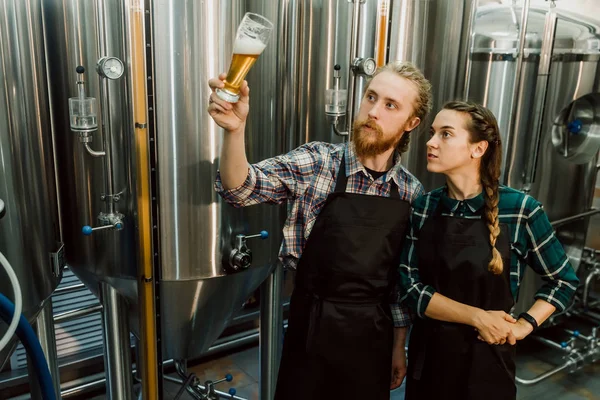 양조장 근로자는 유리 튜브에서 갓 만든 맥주를보고 논의합니다. 양조장 공장에서 맥주를 테스트하는 남성 및 여성 양조장. 4k. 소규모 비즈니스 개념. 스톡 사진