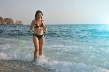 güzel genç kadın sahilde gün batımında sörf boyunca yürüyor.