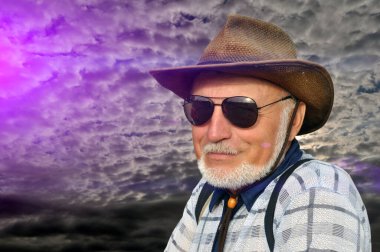 Şapkalı, siyah güneş gözlüklü ve sakallı bir adamın portresi. Gün batımında doğada dinlenirken gülümsemek ve bakmak. Boyama