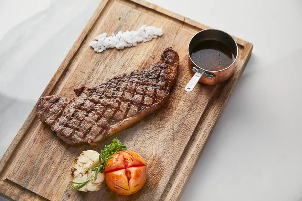 Striploin steak served with roasted vegetables and gravy sauce on cutting board, white marble background