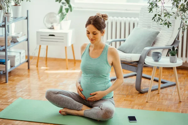 Pregnant young woman sitting on exercise mat and holding hands on her stomach