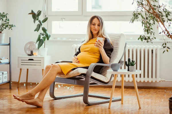 Pregnant young woman sitting in rocking chair, holding hand on belly and using mobile phone
