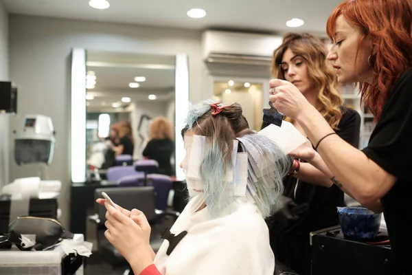 Två unga Frisör färgning hår av kvinnlig klient medan hon läser hennes mobiltelefon — Stockfoto