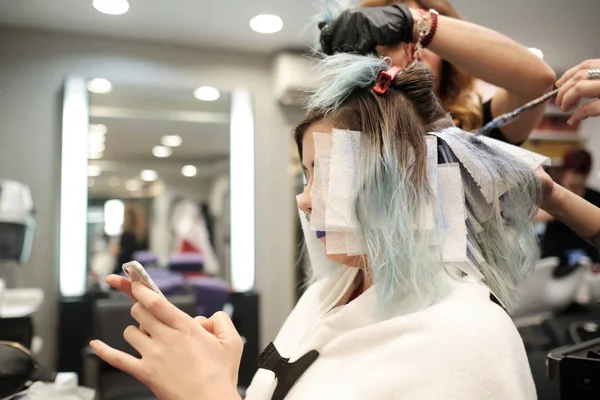 Dos peluquero joven teñir el pelo de cliente femenino mientras lee su móvil —  Fotos de Stock