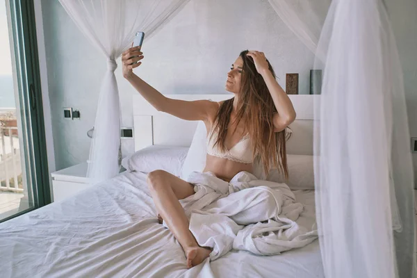 Eine junge Frau in Unterwäsche auf ihrem Bett macht ein Selfie — Stockfoto