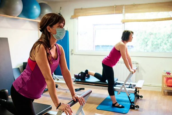 Egzersiz yapan bir grup olgun kadın, sosyal mesafeli Pilates ve Yoga yapıyor ve yüz maskesi takıyor.