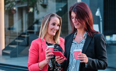 İki iş kadını ofis dışında cep telefonu kullanıyor ve kahve içiyor.