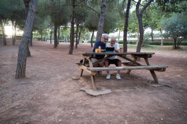 İki emekli adam, sakin bir park ortamında piknik masasında dijital tablet kullanarak boş zamanın tadını çıkarıyor.