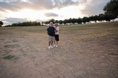 Parkta kucaklaşan yaşlı çift, huzurlu bir günbatımının tadını çıkarıyorlar, emekliliklerinde uzun süreli aşkı ve arkadaşlığı simgeliyorlar.