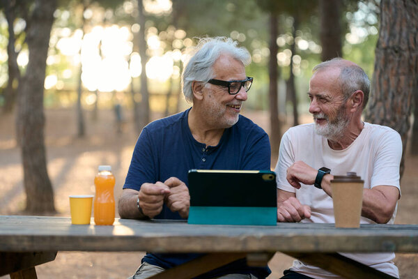 Two retired men enjoying leisure time in nature, using a digital tablet and having a conversation at a picnic table