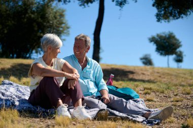 Güneşli bir günün tadını çıkaran gülümseyen emekli çift parkta bir piknik battaniyesinde oturup birlikte eğlenirken kahkaha ve sevgiyi paylaşıyorlar.