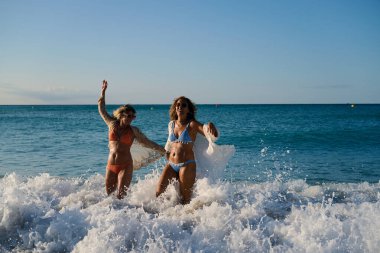 Bikini giyen ve plaj örtüsü takan iki olgun kadın güneşli bir sahilde el ele tutuşup kollarını kaldırarak dalgaların tadını çıkarıyorlar.