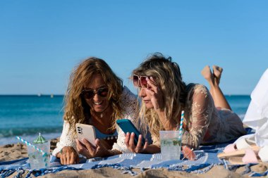 İki olgun kadın plaj havlusunda uzanıyor, kokteyllerin tadını çıkarıyor ve akıllı telefonlara bakıyor, yaz tatilinde eğleniyorlar.