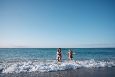 İki kadın kumlu sahilde neşeyle koşuyor, sıcak güneşi kucaklıyor ve okyanusun kenarında mükemmel bir yaz gününde dalgalar sıçrıyor.