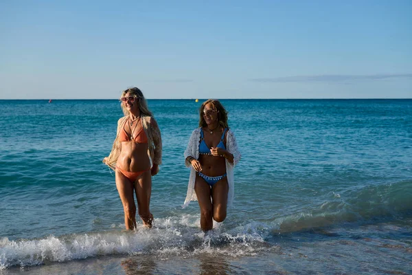 Bikini giyen ve plaj örtüsü takan iki olgun kadın okyanus dalgalarında yürüyüp güneşli bir plaj tatilinin tadını çıkarıyorlar.