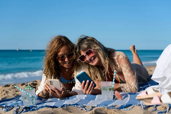 İki gülümseyen kadın plaj havlusunda uzanır, akıllı telefonlar kullanır, güneşli yaz günlerinde ferahlatıcı içkilerin keyfini çıkarırlar.