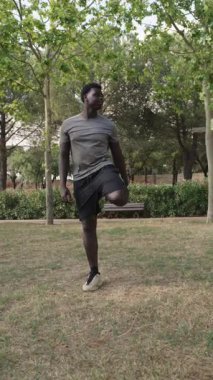 Sakin park ortamında bacaklarını esneten genç atletik erkek, huzurlu yaz antrenmanı sırasında odaklanarak ısınma egzersizi yapıyor.