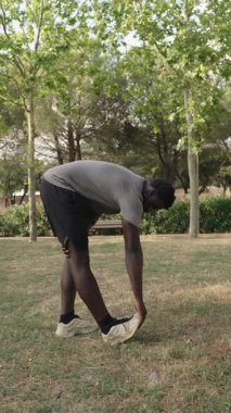 Açık hava fitness rutini sırasında bacak kaslarını esneten atletik siyah erkek, ağaçlar ve çimlerle birlikte güneş ışığında yavaş çekim sekansıyla ısınıyor.