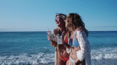 Bikinili iki neşeli olgun kadın, güneşli bir sahilde kokteyl içip, gülüyor ve ağır çekimde kadeh kaldırıyor.