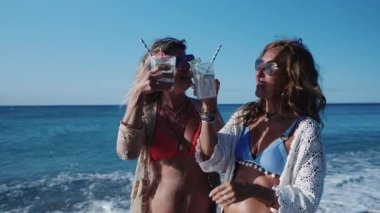 Bikinili iki olgun kadın güneşli bir sahilde kokteyllerini yudumluyor, gülüyor ve ağır çekimde kadeh kaldırıyor.