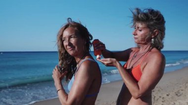 İki olgun kadın kumsalda güneşli bir günün tadını çıkarıyorlar. Yavaş çekim montajında güneş kremi sürüyorlar.