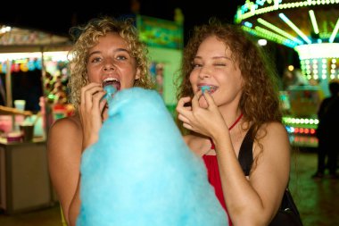 İki arkadaş canlı bir lunaparka gülerken ve gülümserken pofuduk mavi pamuk şekerin tadını çıkarıyorlar. Arka planda renkli bir atlıkarınca parıldıyor.