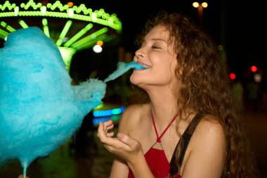 Kıvırcık saçlı genç bir kadın arka planda bulanık bir atlıkarıncayla gece karnavalında gözleri kapalı mavi pamuk şekerin tadını çıkarıyor.
