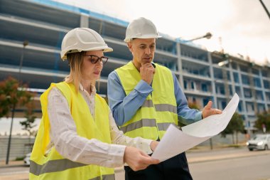 İki inşaat işçisi, inşaat planlarını gözden geçirip inşaat alanındaki işleri tartışmak için çelik yelek giyiyorlar.