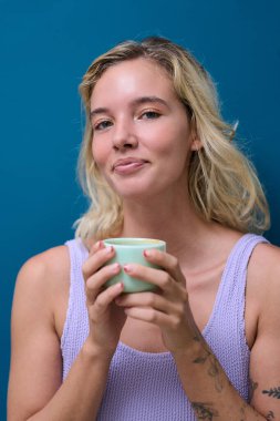 Dövmeli genç bir kadın sıcak bir içkinin tadını çıkarıyor, canlı bir deniz mavisi arkaplanına mutluluk saçıyor, rahat ve rahat bir atmosferi somutlaştırıyor.