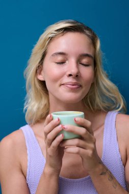 Huzurlu genç bir kadın, altın bir fincan sütün tadını çıkarıyor, gözleri mutlulukla kapalı, canlı mavi bir zemin üzerinde.