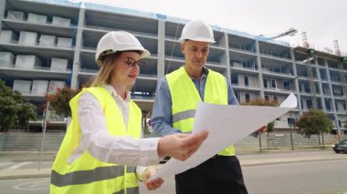 Two professional architects analyzing blueprints together. A man and a woman in hard hats and safety vests discussing the construction project