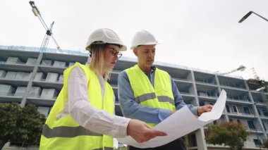 Professional man and woman architect team wearing hard hats and safety vests discussing a building project and examining blueprints on site