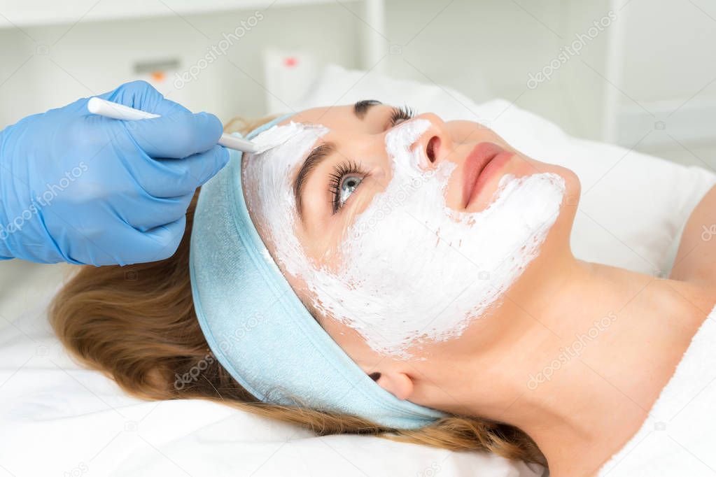 Relaxed young woman is getting facial skin care treatment at beauty salon. Beautician is touching brush with clay to face girl