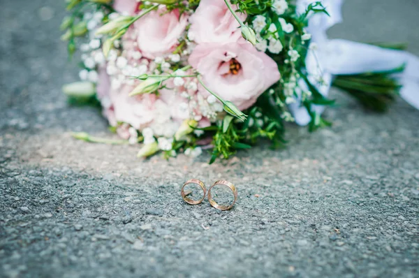 Różowy z zielonym bukiet kwiatów i parę złotych pierścieni leży na asfalcie — Zdjęcie stockowe