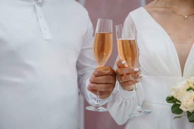 Yeni evli bir çift, beyaz giyinmiş, yumuşak pembe arka planı ve altın köpüklü içkisi olan neşeli bir düğün töreninde şampanya kadehlerini tokuşturur. zarif stil
