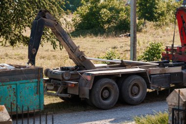 Paslı yeşil bir konteynır gri asfalt yolun yanındaki çimenli bir arazide bulunan hidrolik kollu büyük bir vinç kamyonuna yükleniyor. Sahne endüstriyel temizlik, atık yönetimi ve geri dönüşüm faaliyetlerini iletir
