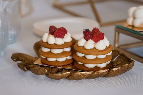 Taze kırmızı ahududu ile kaplanmış ve tüylü beyaz kremayla kaplı iki narin baharatlı kek ustalıkla altın yapraklı bir tepside sunuluyor.