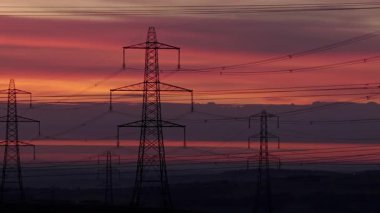 İngiltere 'nin Yorkshire kırsalında güneş doğarken siluetli elektrik direkleri ve elektrik hatları. Yüksek karşıtlık gökyüzü, durağan çerçeve, düz ufuk.