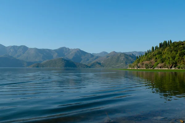 Sabahın erken saatlerinde gölün, ufkun ve arka planda dağların olduğu Skadar Gölü 'nün bir görüntüsü; turistleri taşıyan tekneler bir gezide süzülüyor. Karadağ, Skadar Gölü, 13 Ağustos 2025