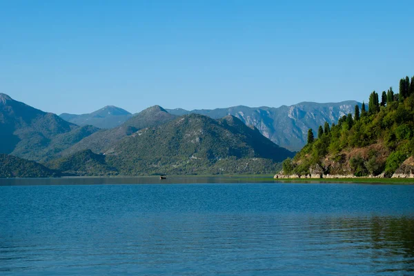 Sabahın erken saatlerinde gölün, ufkun ve arka planda dağların olduğu Skadar Gölü 'nün bir görüntüsü; turistleri taşıyan tekneler bir gezide süzülüyor. Karadağ, Skadar Gölü, 13 Ağustos 2025