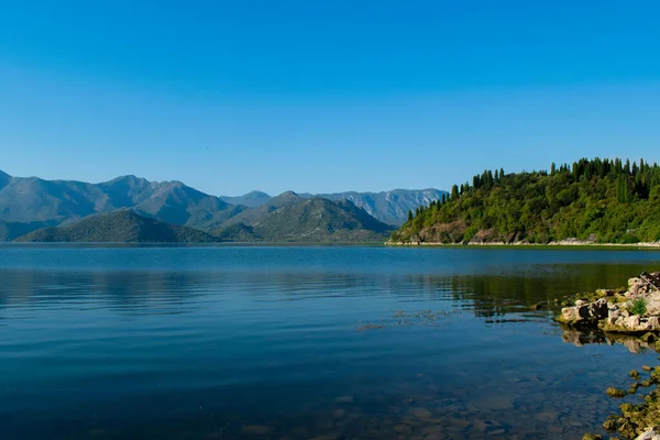 Sabahın erken saatlerinde gölün, ufkun ve arka planda dağların olduğu Skadar Gölü 'nün bir görüntüsü; turistleri taşıyan tekneler bir gezide süzülüyor. Karadağ, Skadar Gölü, 13 Ağustos 2025