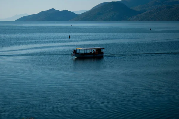 Sabahın erken saatlerinde gölün, ufkun ve arka planda dağların olduğu Skadar Gölü 'nün bir görüntüsü; turistleri taşıyan tekneler bir gezide süzülüyor. Karadağ, Skadar Gölü, 13 Ağustos 2025