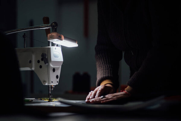 Швейная машина в темноте с рабочими руками на заднем плане
