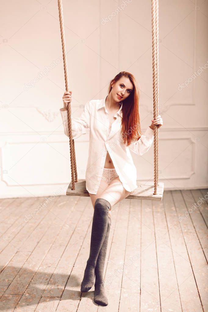 Cute redhead woman with long hair sitting on swing in man s shirt long socks and sexy white lingerie