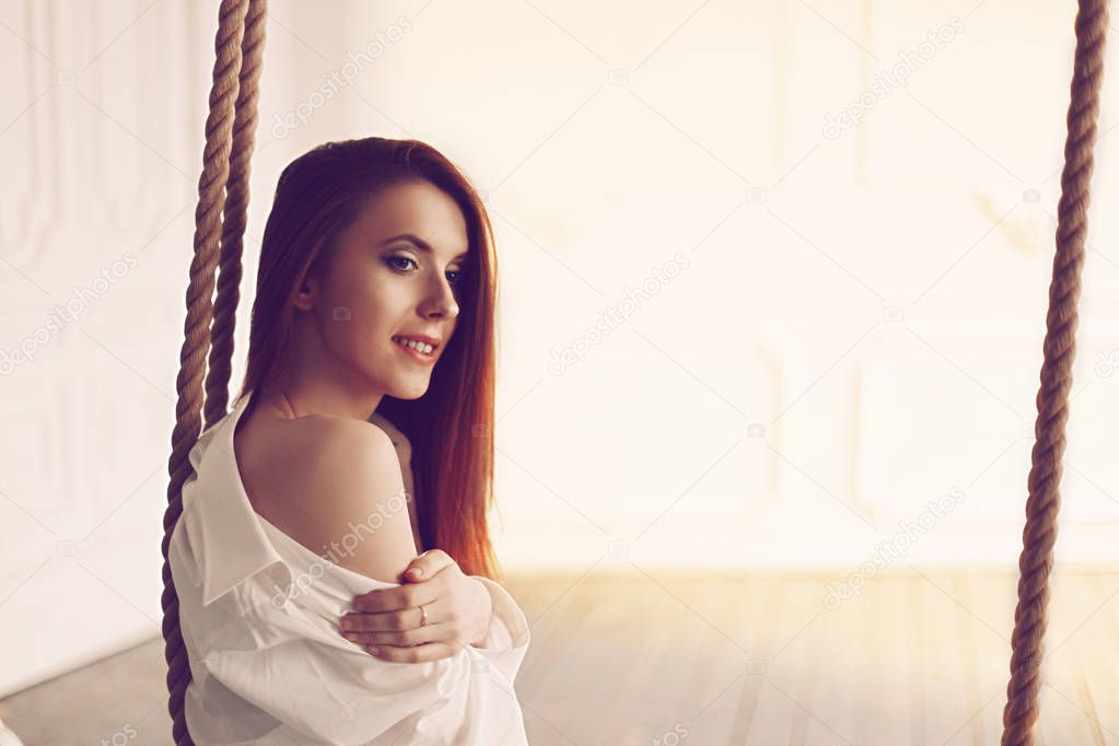 Cute young redhead woman with long hair sitting on swing in man s shirt long socks and sexy white lingerie